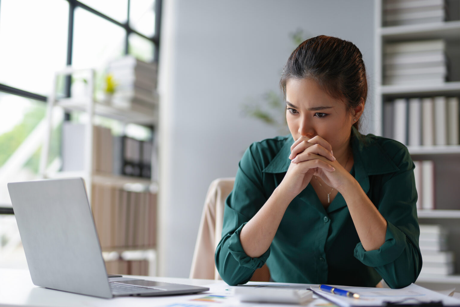 A woman at a desk resting her chin on her clasped hands, looking away from her laptop